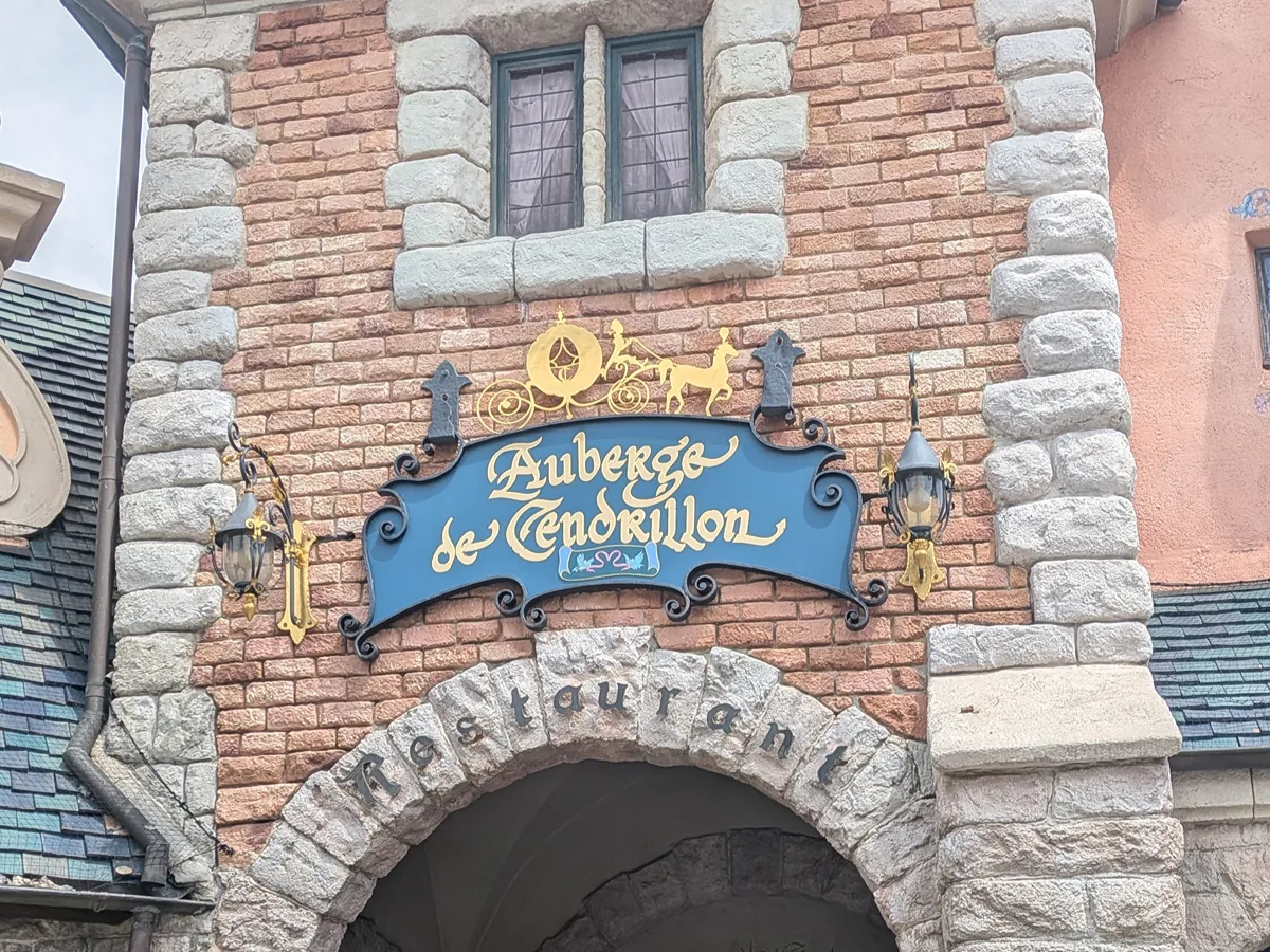 Brick and stone facade of the Auberge de Cendrillon at Disneyland Paris, with the restaurant's blue and gold sign under a green-framed window and the stone arch with the word "Restaurant"