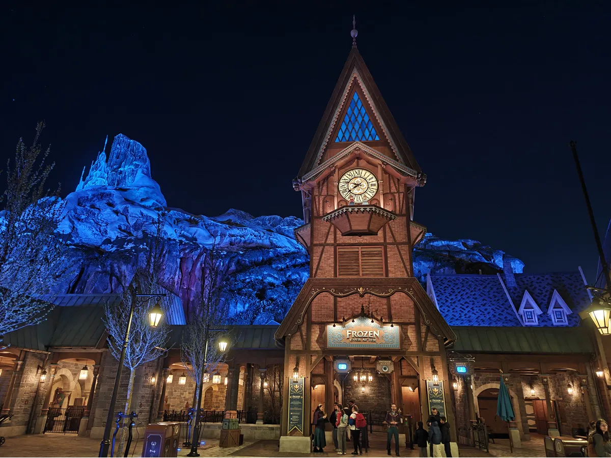 Entrée de Frozen Ever After à World of Frozen, avec la tour de l'horloge illuminée, la montagne en arrière-plan et des visiteurs près des panneaux d'information de l'attraction, de nuit